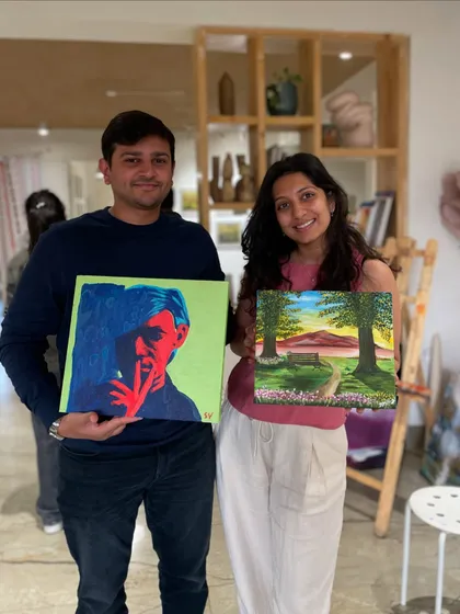 A couple proudly displays their finished paintings. He created a pop-art piece, while she painted a serene landscape, showing the variety of styles you can explore.