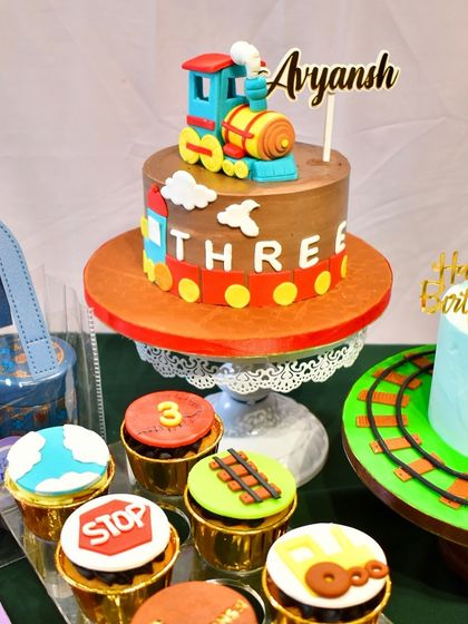A closer look at the train-themed dessert table, showing the main cake with a fondant train topper, a second smaller cake, and a variety of cupcakes with different train-related designs.