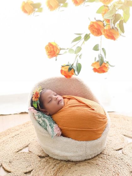 My little sunshine. This simple and sweet portrait features a baby curled up in a cozy chair under a branch of orange roses, creating a warm and gentle image.