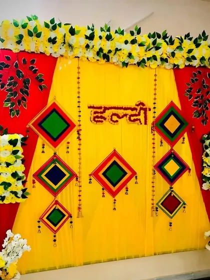 A colorful Haldi backdrop combining red and yellow fabrics for a bold look. The setup is decorated with hanging traditional motifs and bordered with artificial white and yellow flowers, creating a rich and festive frame.