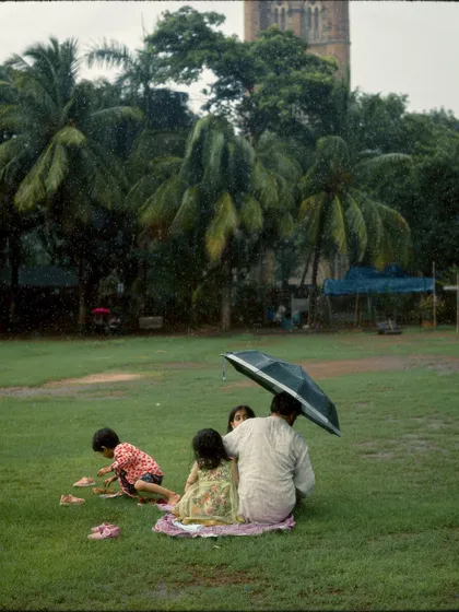 A family enjoys a picnic on the grass at Oval Maidan, huddled under an umbrella in the rain. It's a sweet, intimate moment of finding joy and making memories despite the weather.