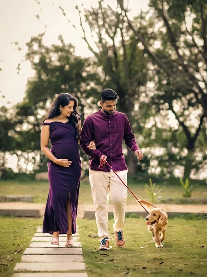 An expecting couple takes a peaceful walk through a park with their dog. The mother-to-be wears an elegant purple off-the-shoulder dress, creating a beautiful family portrait.