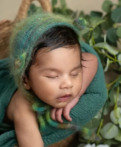 A close-up of a newborn in the chin-on-hands pose, nestled in a basket with a fuzzy green bonnet. The textures and soft light create a dreamy, peaceful image.