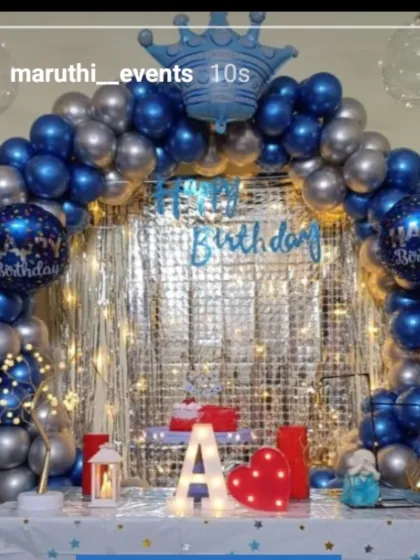 A simple and elegant birthday table setup with a blue and silver balloon arch against a shimmer curtain with fairy lights.