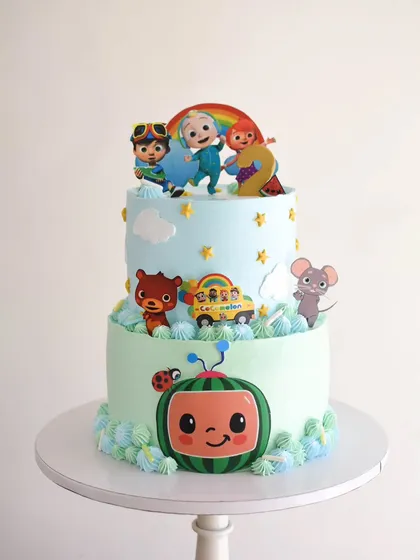 A cheerful two-tiered Cocomelon cake, perfect for a toddler's birthday. The design features edible character cutouts, a soft blue and green color scheme, and playful details like clouds and stars.