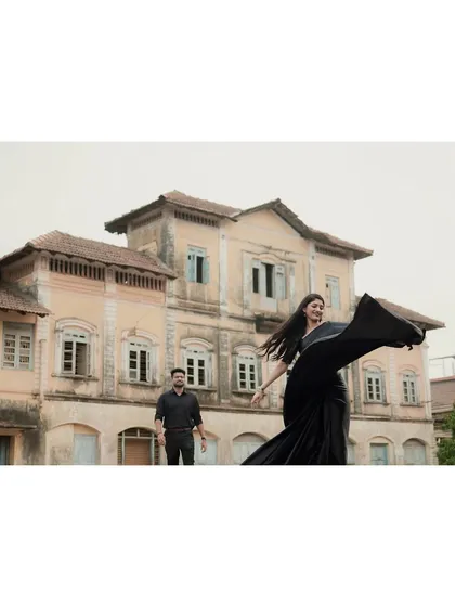 A wide shot showcasing the couple against a grand, old building. The woman's flowing black saree adds a dramatic touch to the scene.
