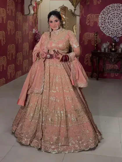 A full-length shot of a client in a peach-pink embroidered lehenga, showing the complete look against a decorative wall.