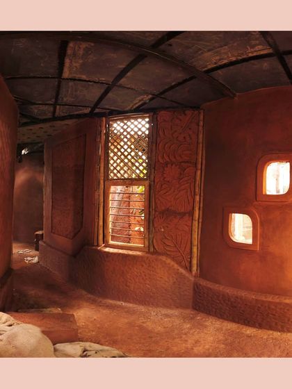 The interior of the cob house reveals its organic form. The undulating mud walls and built-in niches create a cave-like sense of shelter, while a latticed window allows for ventilation and dappled light.