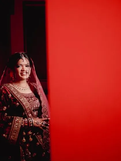 A creative shot using a bold red background to frame the bride. Her traditional maroon velvet lehenga and soft, glowing makeup create a striking contrast.