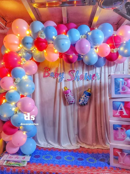 A full view of our pink and blue baby shower balloon decoration. This setup includes a balloon arch woven with fairy lights, a simple curtain backdrop, 'BABY' blocks, and themed foil balloons like the baby bottle.