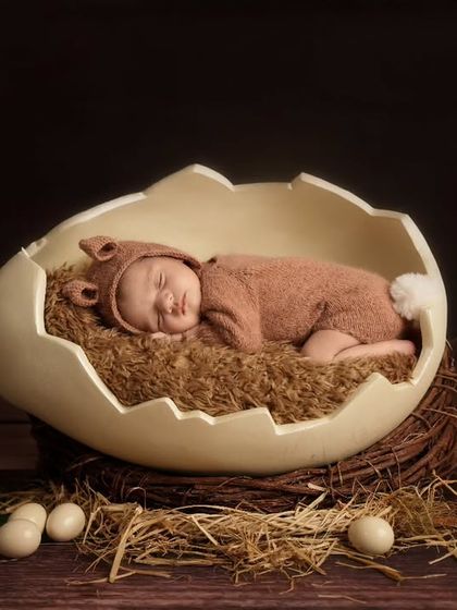 A newborn in a brown bunny outfit sleeps inside a large eggshell prop on a nest, creating a whimsical Easter-themed photo.