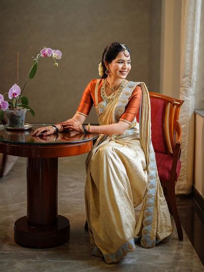 A beautiful, naturally lit portrait of a bride in a classic off-white and rust-orange silk saree. The makeup is kept soft and natural to enhance her features, creating a look of pure elegance.