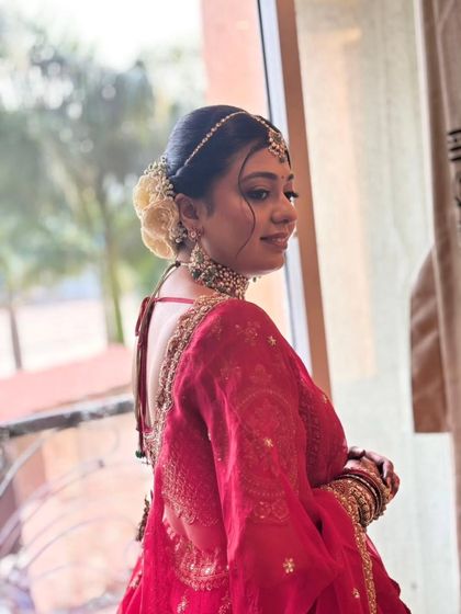 A side profile showcasing the elegant floral bun and the flawless finish of her makeup. The red lehenga and soft lighting create a truly timeless bridal portrait.