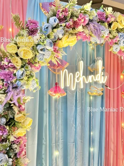A close-up of a pastel Mehendi setup. The arch is filled with beautiful pastel blue, pink, and yellow flowers, with a 'Mehendi' neon sign and cute hanging props.