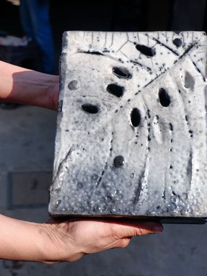 This sculptural tile, held by a workshop participant, shows the dramatic crackle and smoky effects achieved through Raku firing. The holes and textures interact with the unpredictable glaze.