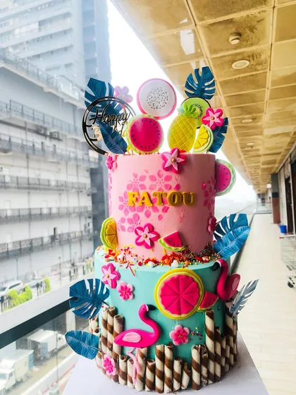 A vibrant and fun two-tiered tropical flamingo cake that's perfect for a summer party. This eggless designer cake is decorated with colorful fondant fruits, tropical leaves, and cheerful flamingos, all wrapped with chocolate wafer sticks.