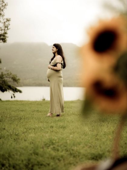 An artistic portrait using a foreground blur of sunflowers to frame the mom-to-be by the lake. It creates a dreamy, painterly effect.