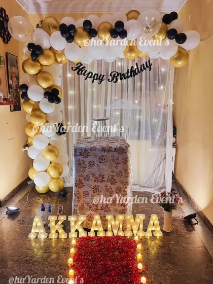 A cozy and romantic birthday surprise at home. This setup features a path of rose petals, marquee letters, and a balloon arch against a fairy-lit curtain.