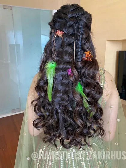 A creative half-up style with multiple braids and colorful feather and floral accessories, perfect for a unique Mehendi look.