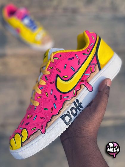 A side view of the pink donut sneaker, showing the dripping frosting effect and the iconic "D'oh" quote written on the midsole. The Nike swoosh is colored to match the yellow of the Simpsons' characters.