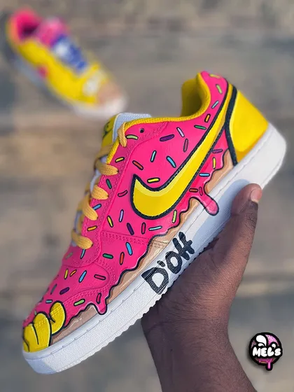 A side view of the pink donut sneaker, showing the dripping frosting effect and the iconic "D'oh" quote written on the midsole. The Nike swoosh is colored to match the yellow of the Simpsons' characters.
