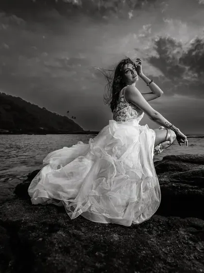 A dramatic black and white bridal portrait by the sea. The moody sky and the texture of the rocks contrast beautifully with the soft, flowing wedding dress, creating a powerful and timeless image.