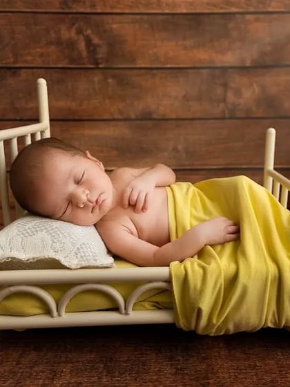 A newborn sleeps on their side in a miniature bed prop against a rustic wood background, demonstrating a classic posed newborn setup.