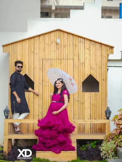 A fashionable portrait of a couple posing in front of a rustic wooden cabin, with the mother-to-be holding a paper umbrella.
