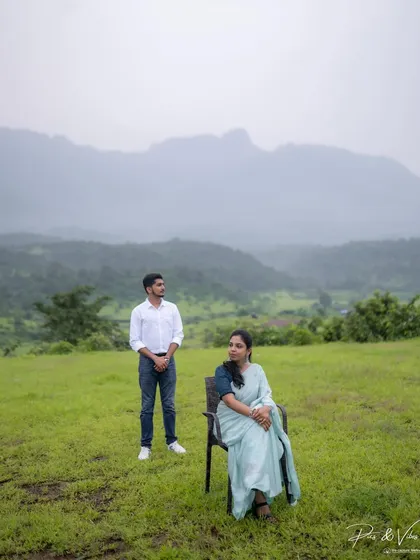 A composed, artistic shot with the bride seated and the groom standing, set against a backdrop of rolling green hills.