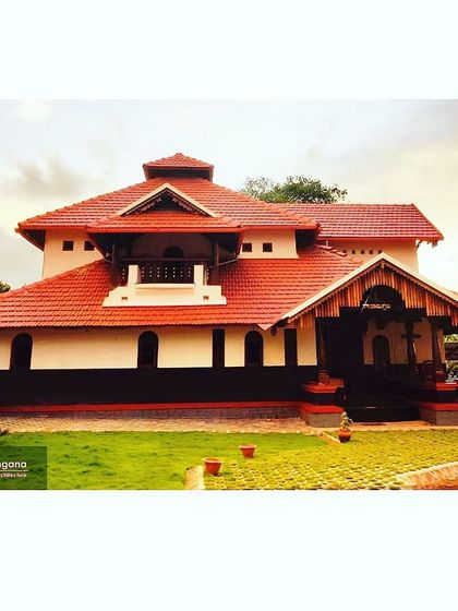 An exterior view of our traditional eco-friendly bungalow in Udupi. The design features a multi-level sloping roof with Mangalore tiles, a prominent balcony, and a lush green lawn, creating a home that is in harmony with its coastal surroundings.