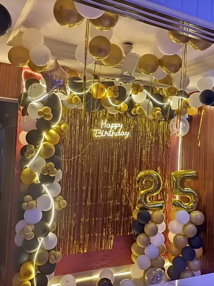 A complete room decoration for a 25th birthday. A gold fringe backdrop is framed with a black, white, and gold balloon arch with neon lights.