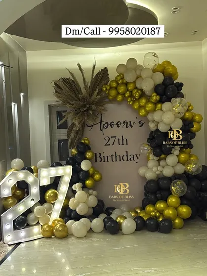 A bold and glamorous 27th birthday setup. The decor features a personalized backdrop framed by a dramatic arrangement of black, white, and gold balloons, perfectly complemented by large light-up marquee numbers.