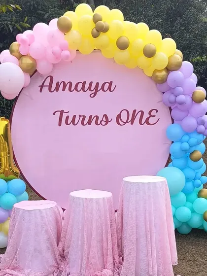 An outdoor first birthday celebration with a custom pink backdrop. The setup is framed by a colorful pastel balloon arch and includes a gold foil number '1' balloon and covered tables for the cake.