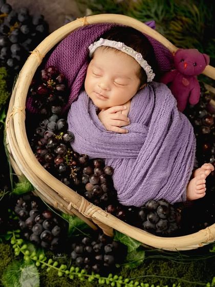 Sweet as a grape. This creative newborn shoot features a baby swaddled in purple, nestled among fresh grapes in a basket for a unique and colorful portrait.