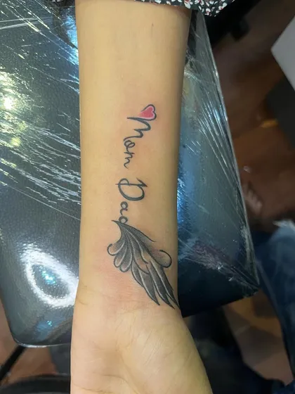 A delicate "Mom Dad" tattoo on the wrist, with a small heart and a single wing. This is a more subtle but equally meaningful tribute.