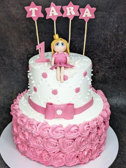 A lovely two-tier princess cake for a first birthday. This crunchy hazelnut cake is decorated with pink rosettes and a handmade fondant doll topper.