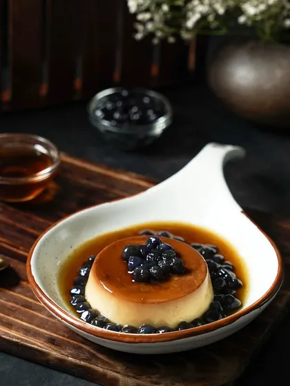 A caramel flan dessert topped with boba pearls, served in a pool of syrup. The dark, woody setting and focused light create a rich and decadent mood for this unique dessert-drink fusion.