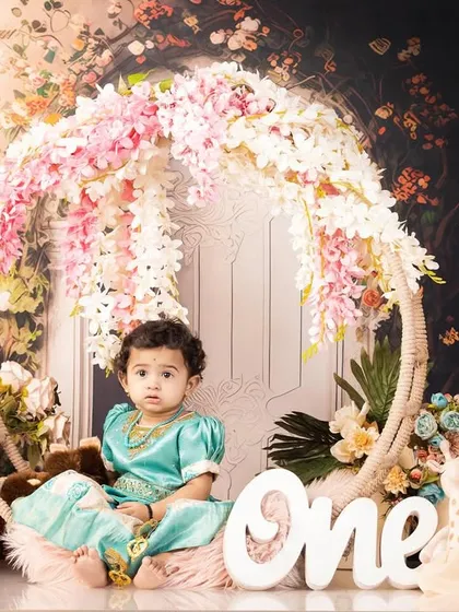 A first birthday portrait of a little girl in a turquoise dress, sitting in a floral hoop swing.