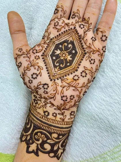 A beautiful party henna design with a central geometric floral motif. The palm is surrounded by delicate leafy vines, and the wrist has a bold, dark cuff.