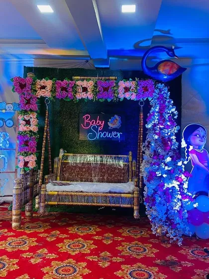 A Radha-Krishna themed baby shower stage setup in a home, complete with a decorated swing, green backdrop, and character cutouts.