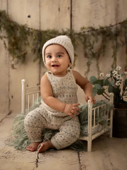 A baby boy in a knit outfit sits on a miniature bed in a rustic, woodland-themed studio setup.