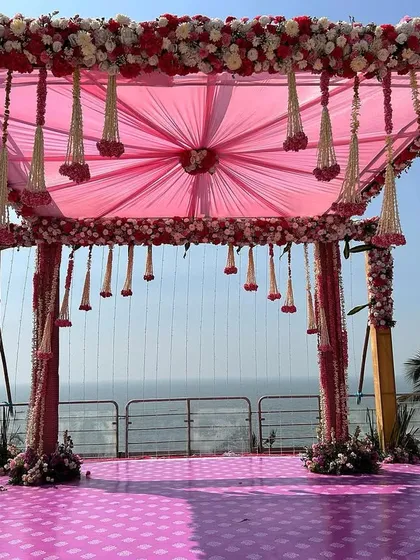 A stunning beachside Vidhi Mandap with pink floral pillars and hanging kaleeras. The design perfectly frames the ocean view, making it a breathtaking location to get married.