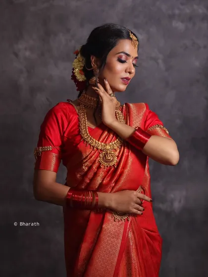 A three-quarter shot of a model in a classic red bridal saree. The pose highlights the neckline of the blouse and the layers of traditional gold necklaces.