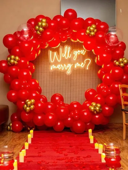 The perfect proposal setup. This decoration features a large ring of red balloons, a 'Will you marry me?' neon sign, and a red carpet aisle lined with candles, creating an unforgettable moment.