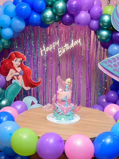 An underwater adventure with a magical mermaid theme. The decor is set with Ariel cutouts, a mermaid tail, and a sea of purple, blue, and green balloons.