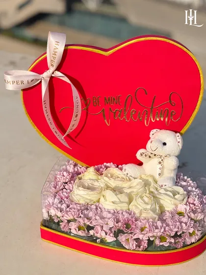 A close-up of the heart-shaped floral hamper, showing the soft teddy bear nestled among the white and purple flowers. It's a sweet and cuddly gift of love.