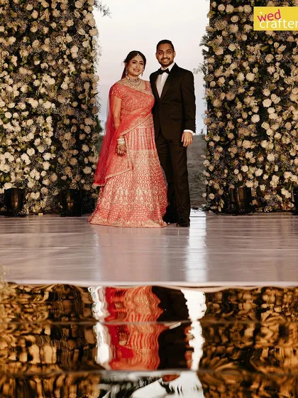 A creative shot of the couple with their reflection in the water, standing between two beautiful white floral walls at their beach wedding.