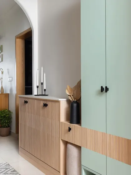 The foyer of 'The Pastel Abode' features a pastel green cabinet paired with oakwood finishes and black accents. The grooved details on the shoe cabinet add a subtle texture to the welcoming space.