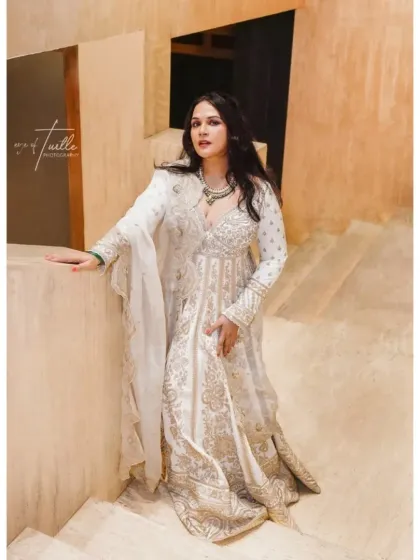 A full-length portrait of the bride, leaning against a modern, minimalist staircase. The neutral background makes her white and gold outfit stand out beautifully.