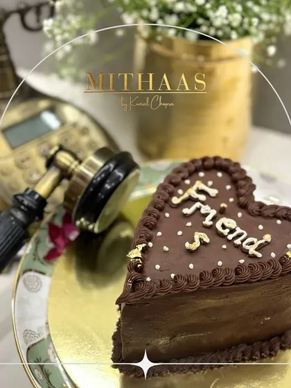A heart-shaped chocolate cake designed to celebrate friendship. The rich frosting is decorated with a piped border, delicate sprinkles, and the word "Friends" written in elegant script.